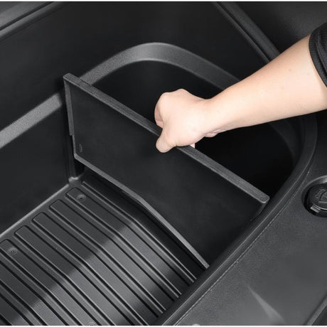 Hånd der placerer sort plastikdeler i den tomme frunk på Tesla Model Y, viser montering af skillerum